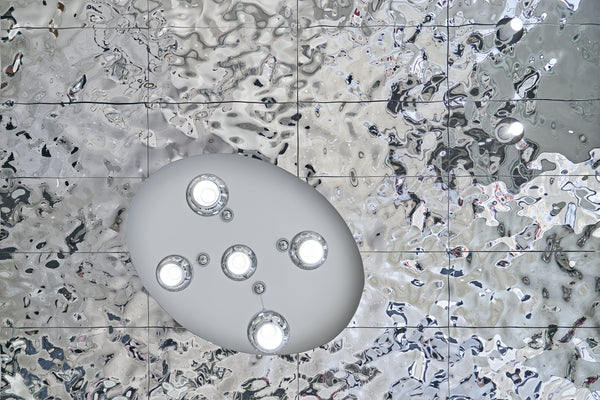 Close-up of Aqua High Wave silver glass panels on a ceiling, reflecting modern spherical pendant lights and creating a fluid, wavy metallic effect.