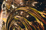 Close-up of a reflective gold glass wall panel with distorted reflections of lights and surroundings in a wavy pattern.