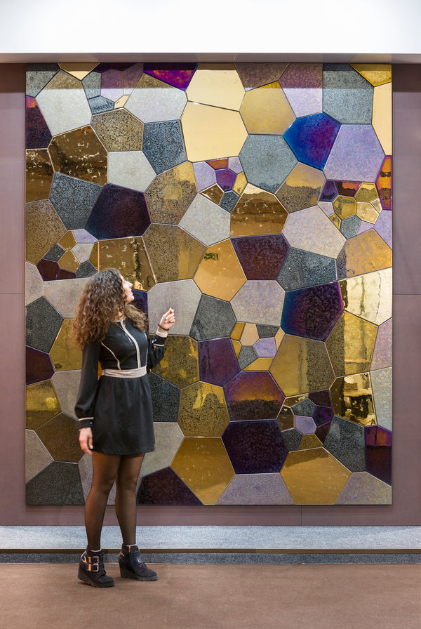 A woman standing in front of a large polygonal mosaic wall panel at an exhibition. The glass textures and metallic hues create a captivating display.
