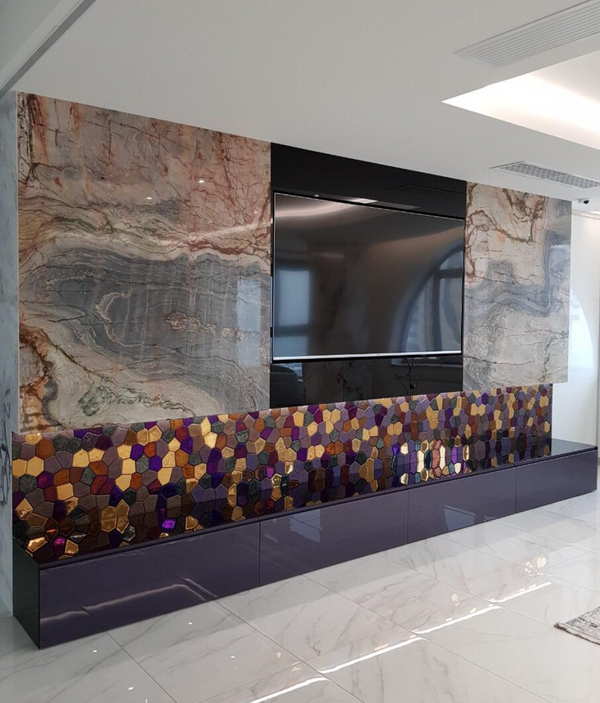 A polygonal glass wall panel placed under a TV in a contemporary interior. The mix of gold, bronze, and iridescent tiles enhances the space.