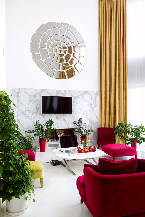 Spiral mirror wall art in a modern workspace with a TV. Marble accent wall, red velvet seating, gold curtains and lush green plants.

