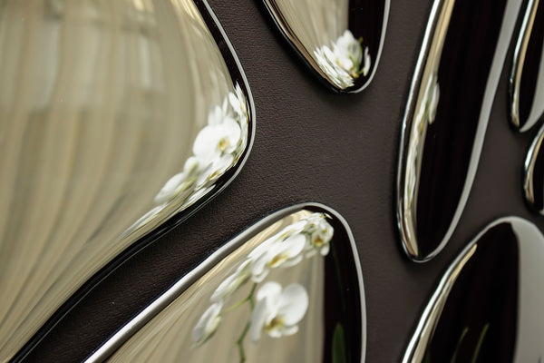 Close-up of modern mirror wall decor with chrome-finished irregular shapes reflecting white orchid flowers and soft curtain folds
